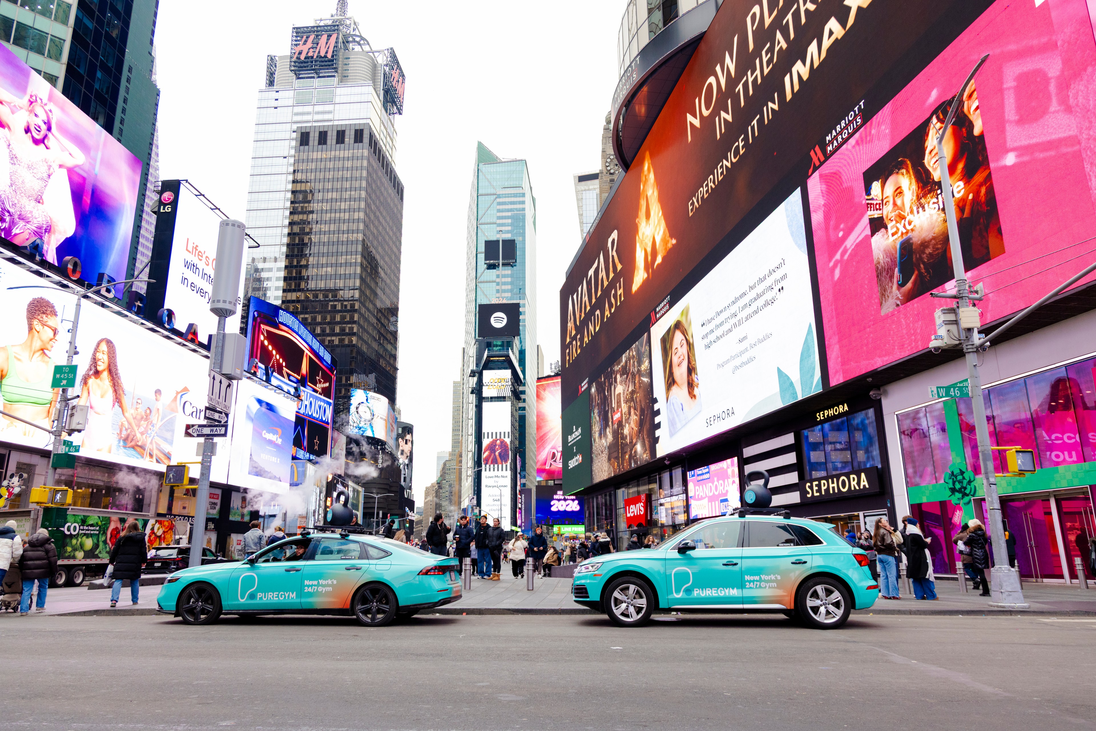 A teal-wrapped SUV branded with the PureGym logo and the text "New York's 24/7 Gym" drives along a city street, with a large black kettlebell prop mounted on the roof rack.