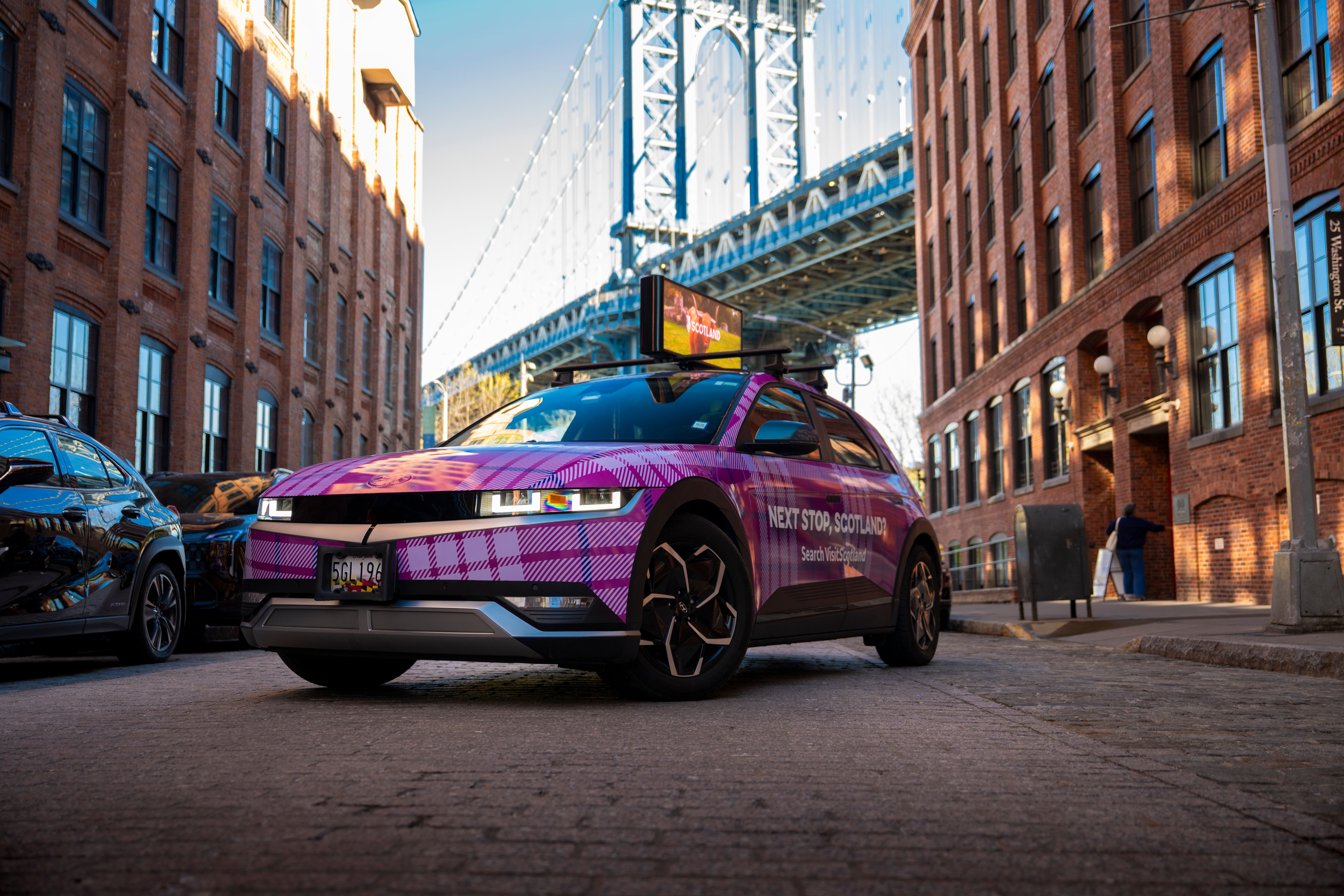 A Hyundai IONIQ 5 wrapped in a bold pink and purple tartan pattern, parked on a cobblestone street between brick buildings. The side of the car reads "Next Stop, Scotland?