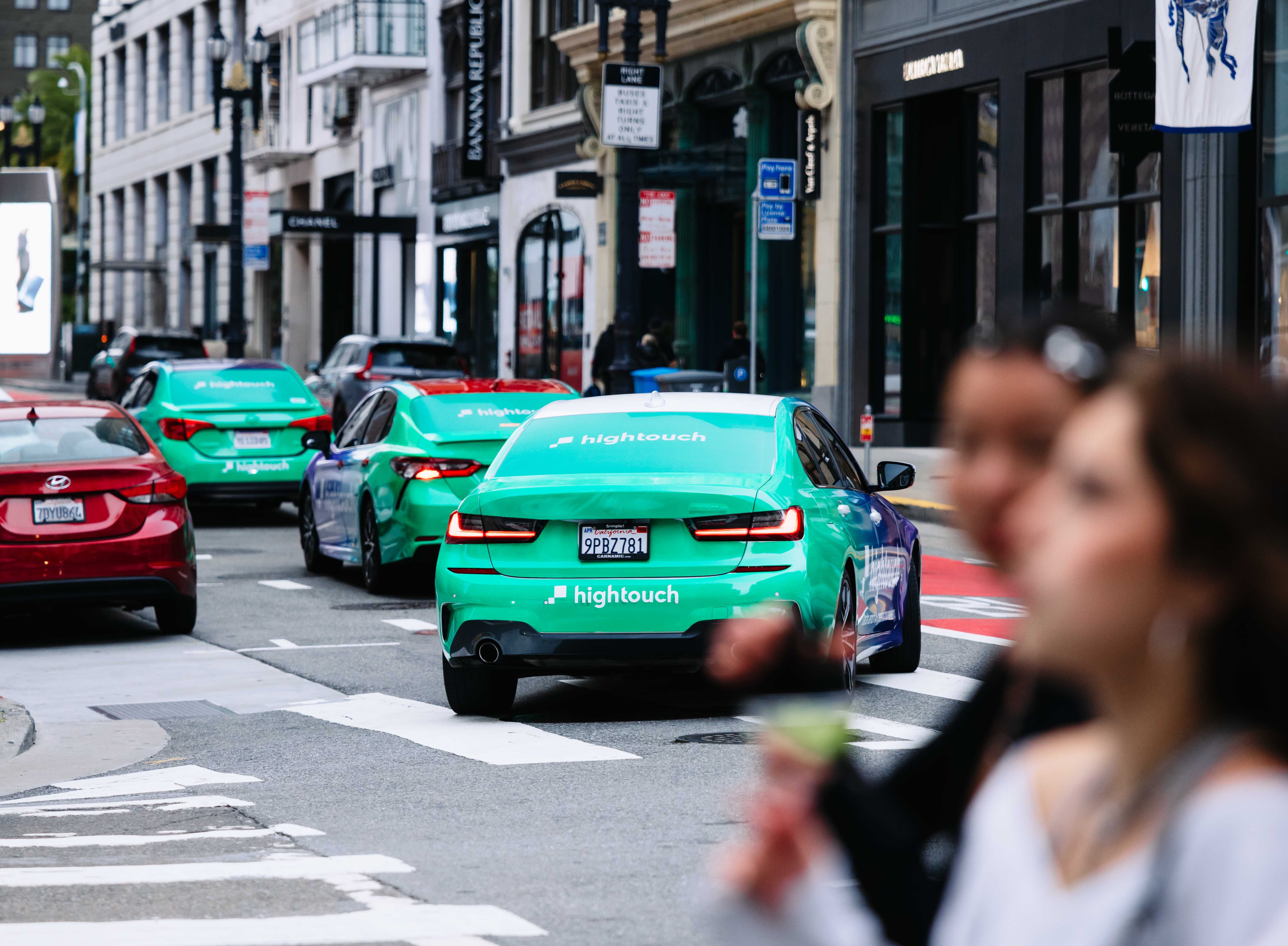 Multiple teal green BMW sedans wrapped in Hightouch branding driving through a busy urban intersection, with pedestrians in the foreground and upscale retail storefronts including Banana Republic lining the street in the background.