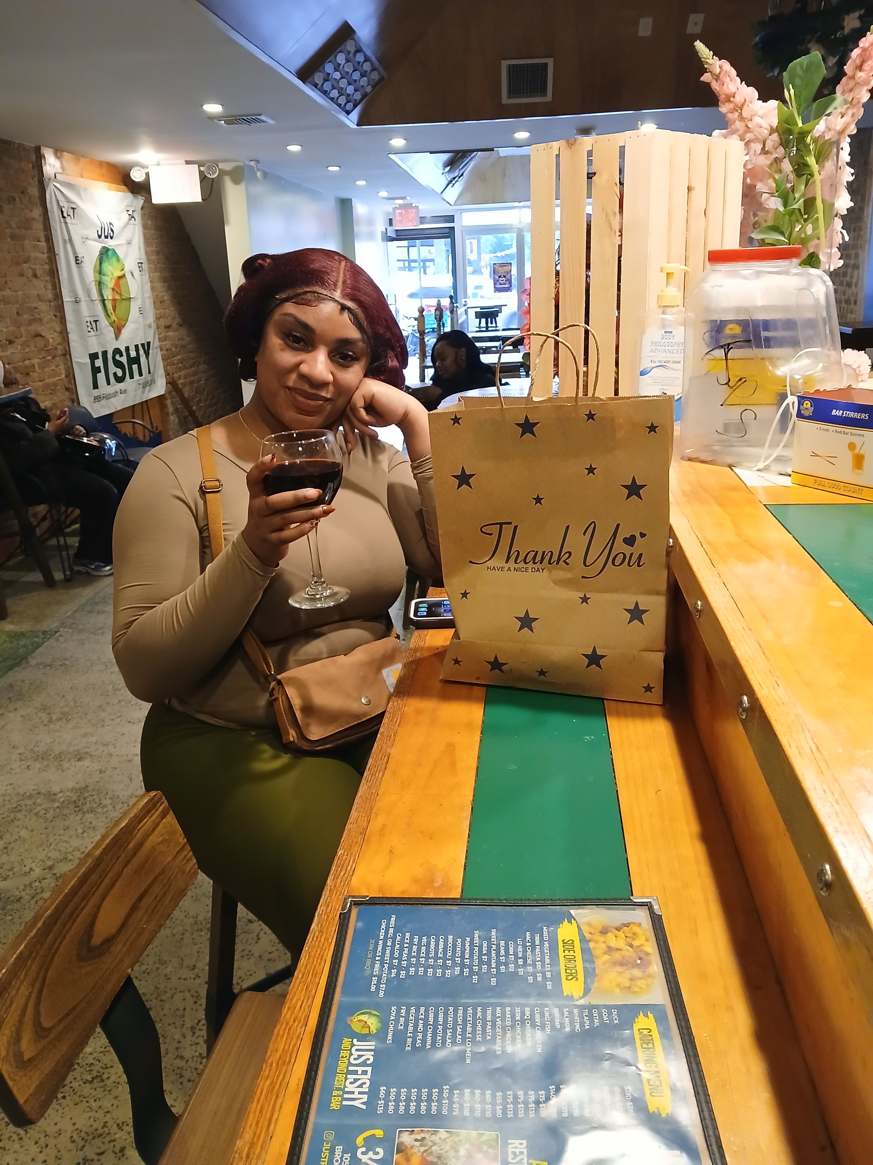 Vibrant bar atmosphere at Jus Fishy & Beyond in Brooklyn, showing a customer with a "Thank You" takeout bag and house wine.
