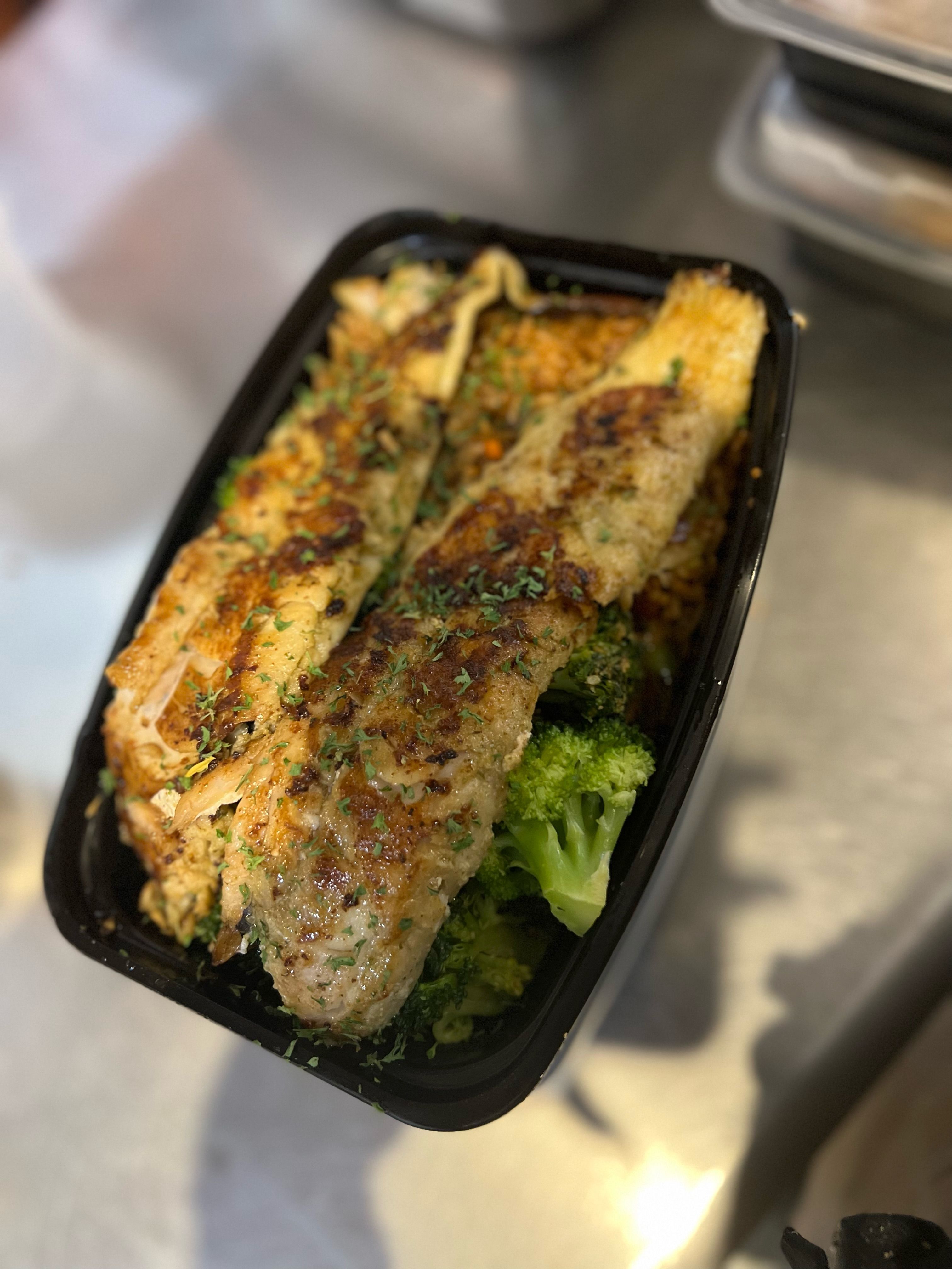 Healthy grilled fish fillet seasoned with island spices, served over steamed broccoli and rice in a takeout container.