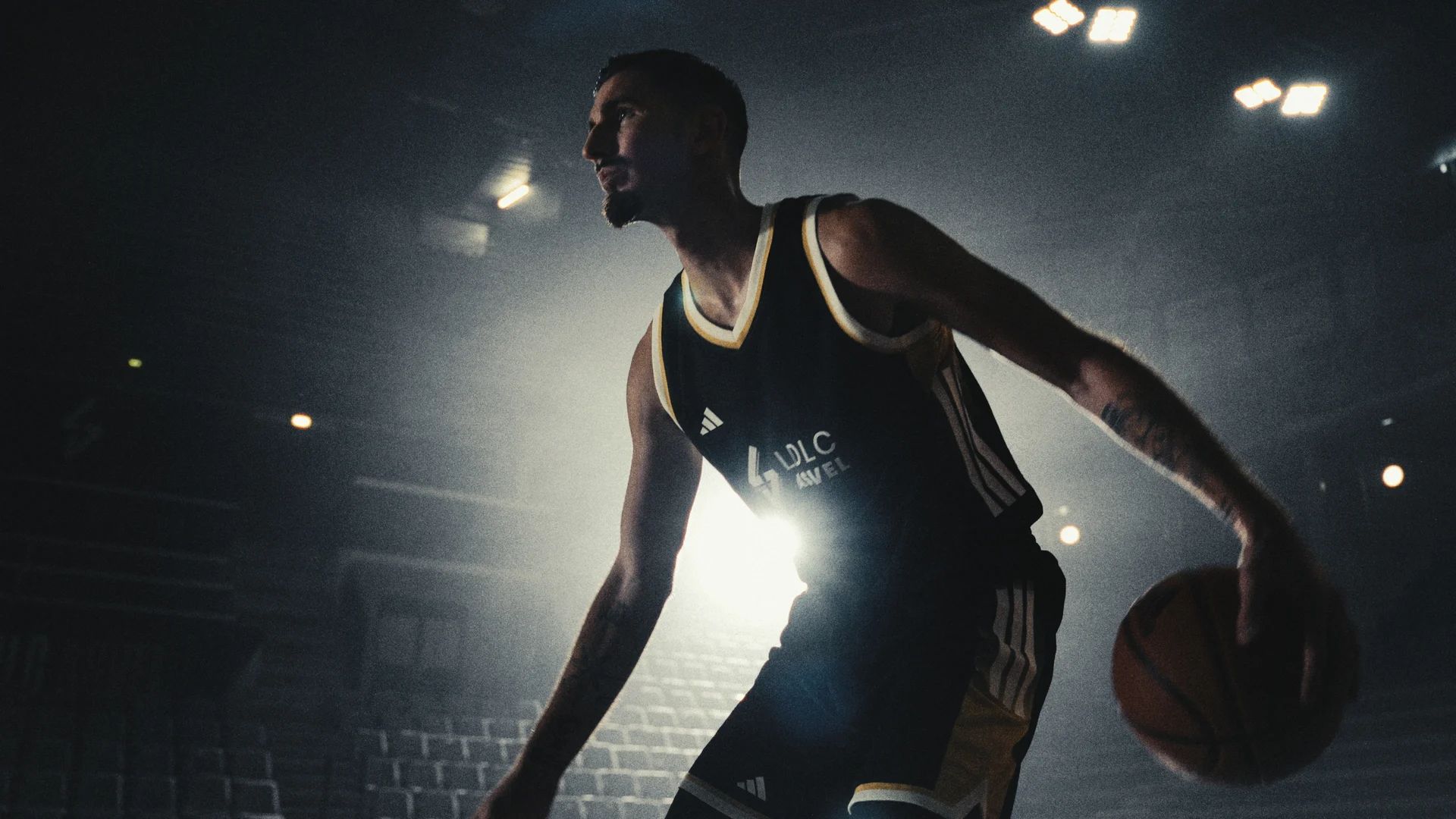 Un joueur dribble en maillot noir et or, devant les lumières de la salle.