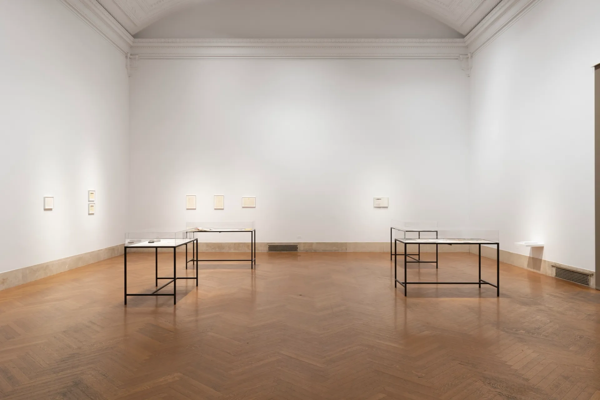 A gallery with a high ceiling and white walls with four vitrines and art lining the walls.