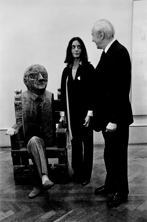 A man and woman stand next to a wooden sculpture of a seated man in a galler
