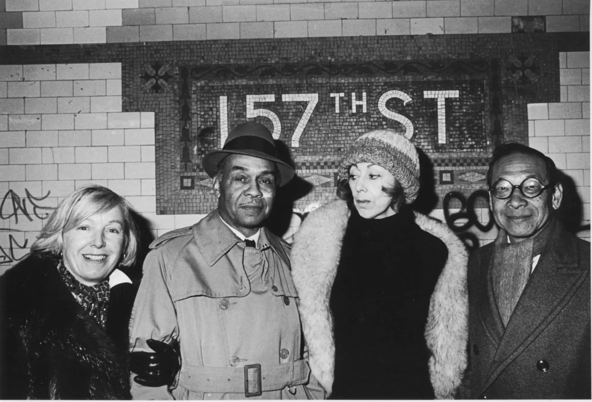 A black and white photograph of four people dressed in coats posing beneath a subway mosaic displaying "157th Street." Graffiti is visible on the surrounding subway tiles.