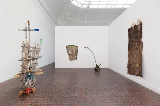 In an enclosed gallery space, two assemblage sculptures made from various everyday materials rest on the floor. A fabric-like work made from natural materials hangs from both of the adjacent walls.