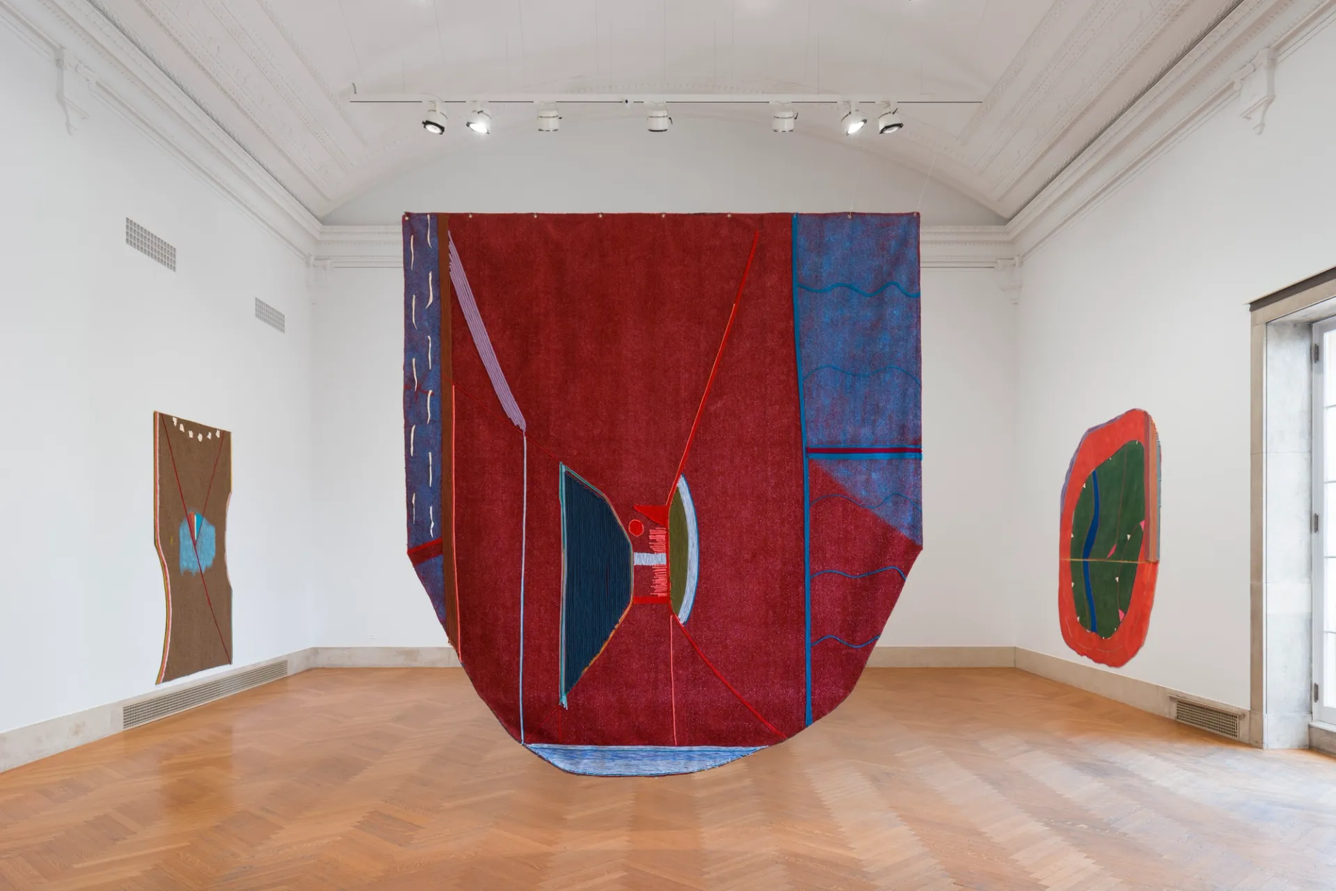 large shield-shaped painting suspended in space in gallery with two large works on the walls on either side visible behind it