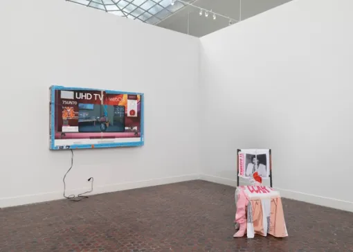 On a wall to the left, a wide flat screen TV with a bright blue frame hangs mounted on a wall. To the right, an altar-like sculpture sits on the floor.
