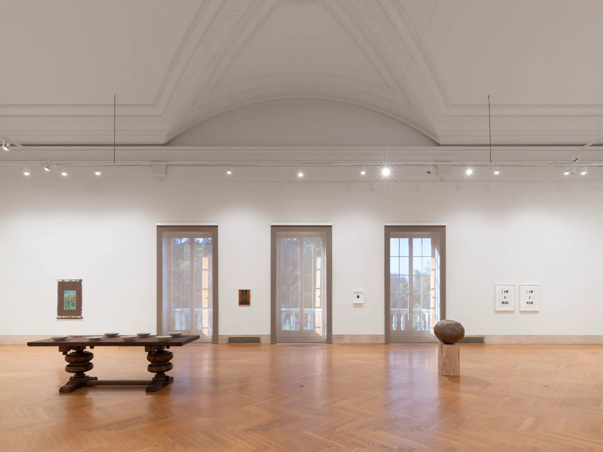 A large enclosed gallery space with three parallel windows, a sculpture and framed artworks to the right, and a carved wooden table holding objects in front of a single framed artwork positioned to the left