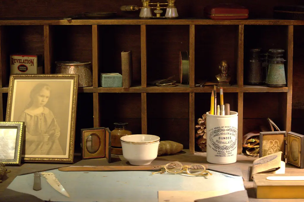 A wooden work desk with a framed sepia portrait, a cup of writing tools, a small bowl, and various containers inside wooden shelves.