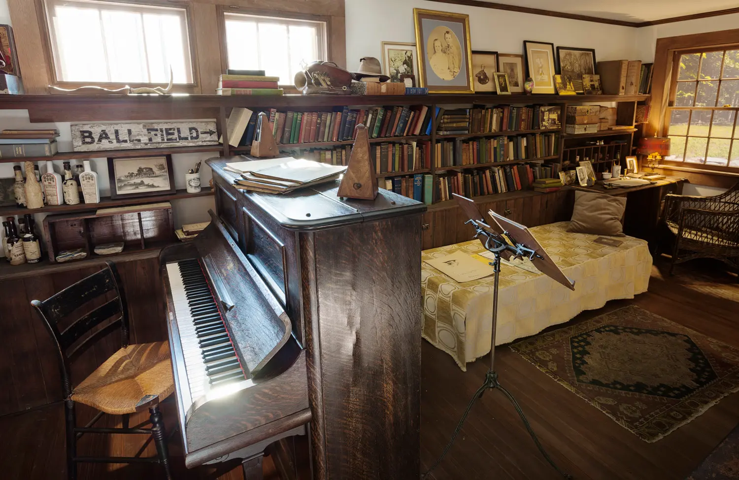 An angled view of a long room shows a piano and chair, a small bed and rug, framed portraits, a bookshelf, and miscellaneous items.