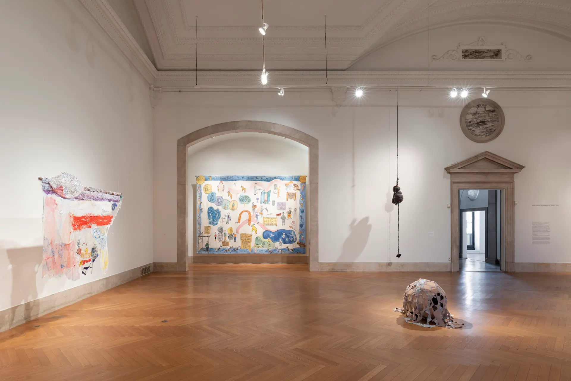 A small abstract sculpture sits on the floor of the center of a gallery. Against a wall to the left, a painting on white, irregular fabric is suspended from the ceiling. Straight ahead is a large painting framed by an arched stone entryway and an abstract sculpture suspended from the ceiling.