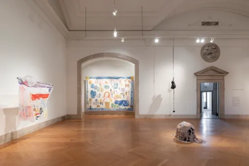 A small abstract sculpture sits on the floor of the center of a gallery. Against a wall to the left, a painting on white, irregular fabric is suspended from the ceiling. Straight ahead is a large painting framed by an arched stone entryway and an abstract sculpture suspended from the ceiling.