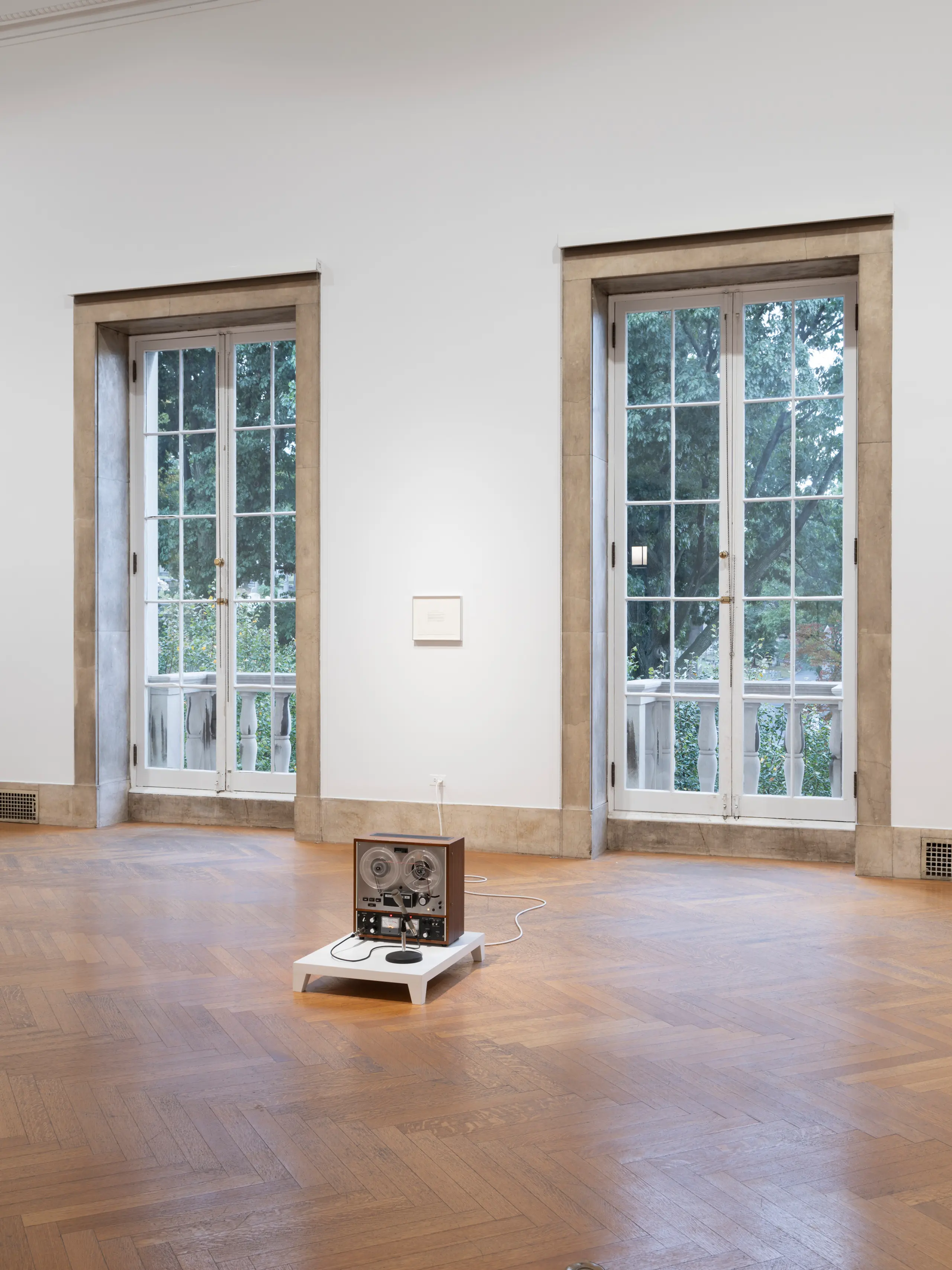 An artwork consisting of a recording device sitting on the floor of a gallery with two windows behind it.