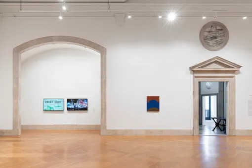 To the left, an arched stone entryway. Directly inside the arched recess, two video monitors displaying video works are positioned on the wall. On the wall to the right is a painting. A lobby with table and another brightly lit gallery beyond are visible through a doorway to the far right.