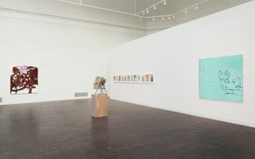In a gallery with Spanish tiled floors, a painting with maroon and pink abstract forms hangs on the left wall. On the right, a series of small colorful works are mounted on the wall beside a bright, teal painting. At the center of the gallery, a small sculpture stands on a wooden pedestal.