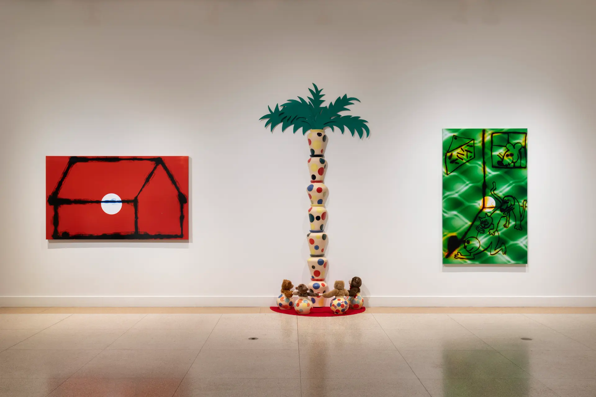 Three artworks are positioned along a gallery wall.  On the left, a rectangular painting with a red background displays the outlines of a house. A painting of cartoon-like images with a green, patterned background hangs on the right. Between these two paintings is a colorful sculpture showing a palm tree and small animals at its base.
