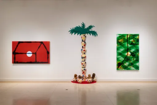 Three artworks are positioned along a gallery wall.  On the left, a rectangular painting with a red background displays the outlines of a house. A painting of cartoon-like images with a green, patterned background hangs on the right. Between these two paintings is a colorful sculpture showing a palm tree and small animals at its base.