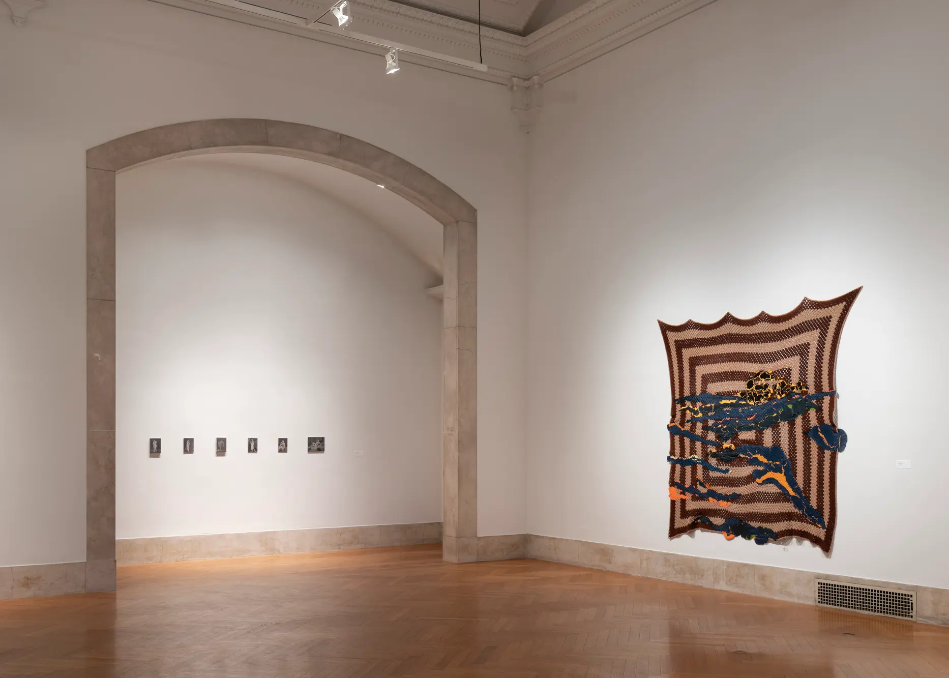 In a gallery, a series of small artworks positioned in a straight line adorn a wall within a large arched entryway. To the right of the entryway, a geometric fabric work hangs on a wall. 