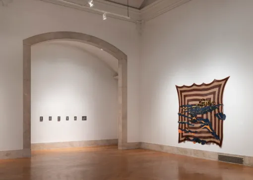 In a gallery, a series of small artworks positioned in a straight line adorn a wall within a large arched entryway. To the right of the entryway, a geometric fabric work hangs on a wall. 