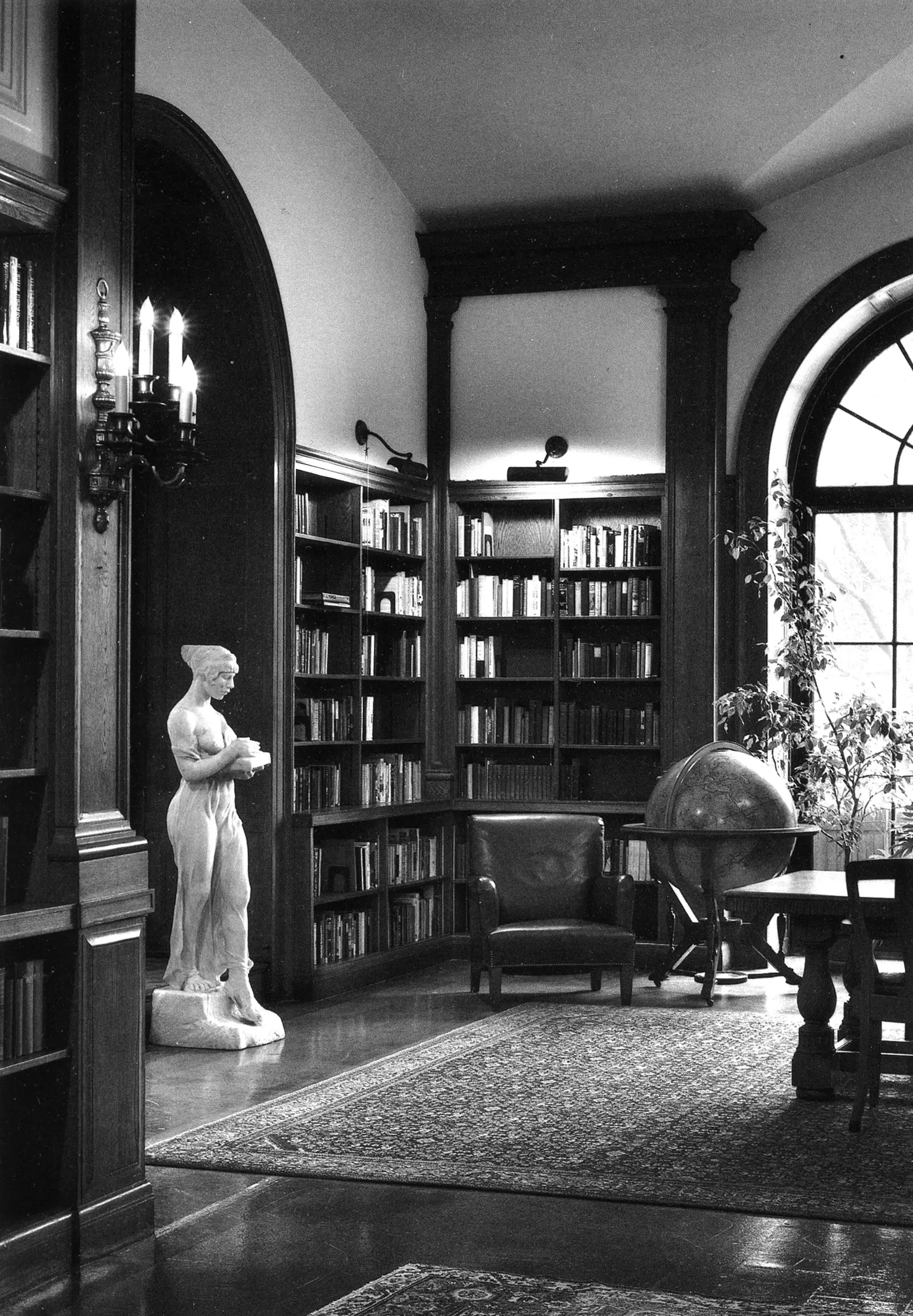 A black and white photograph of a room. To the left, a marble statue of a woman stands beside a bookcase. To the right, a chair and large globe are pictured beside a window.