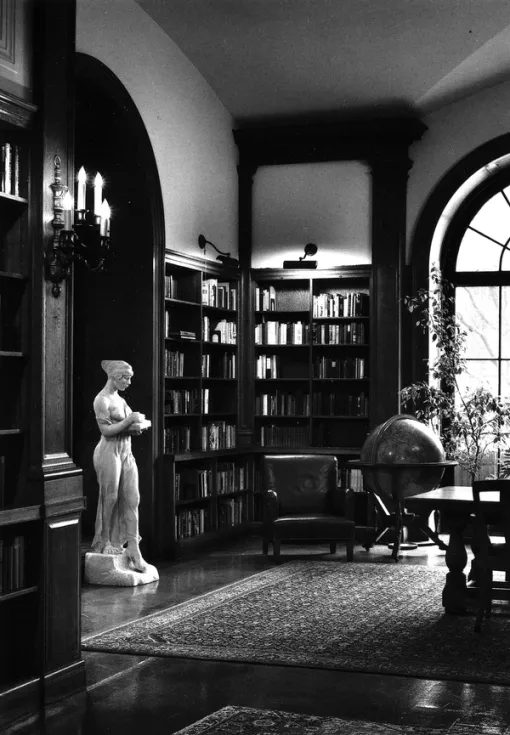 A black and white photograph of a room. To the left, a marble statue of a woman stands beside a bookcase. To the right, a chair and large globe are pictured beside a window.