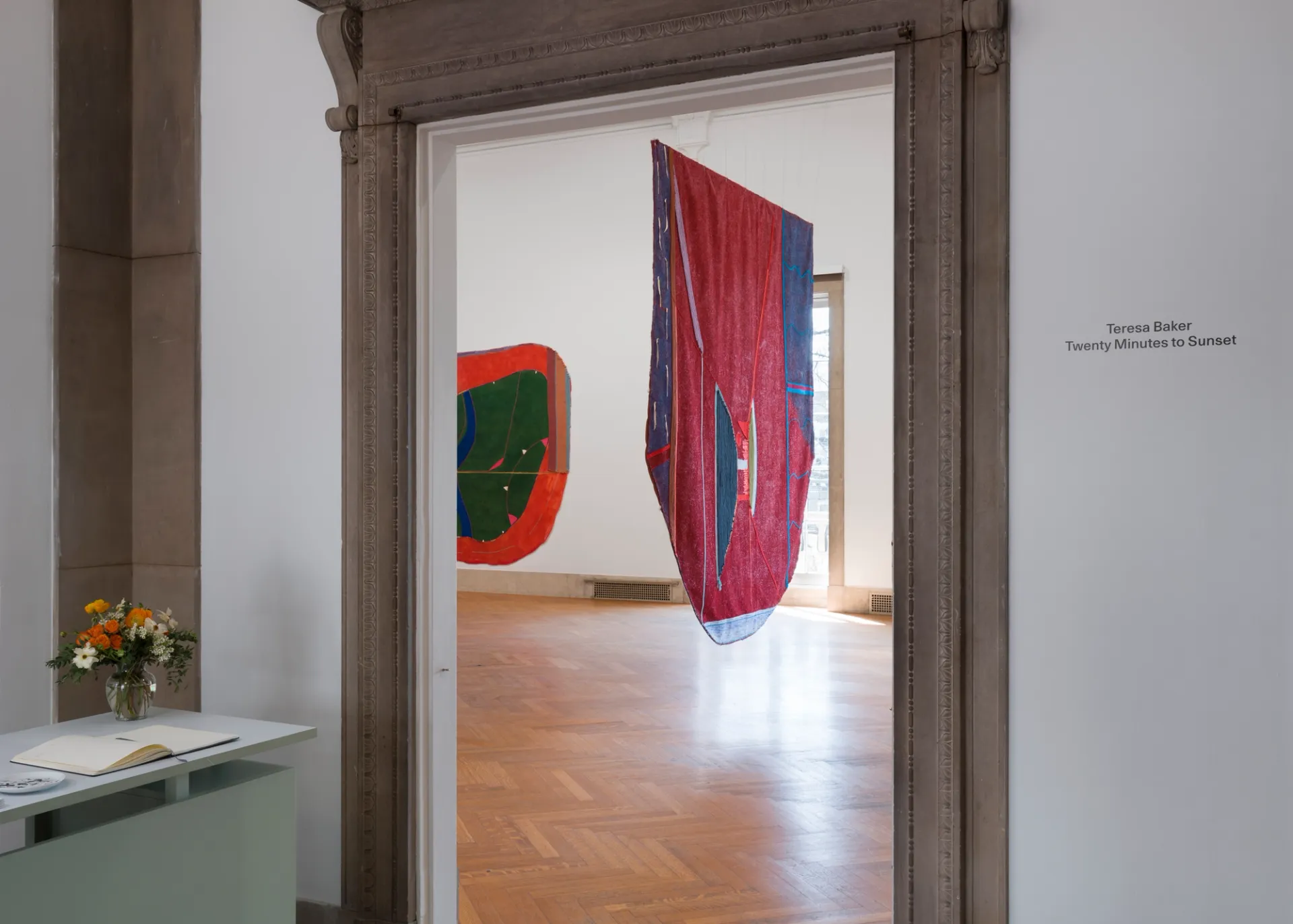 View through a doorway into the gallery; two large paintings visible inside; a reception desk with flowers and a notebook on the left of the door; exhibition title "Teresa Baker Twenty Minutes to Sunset" on the wall to the right