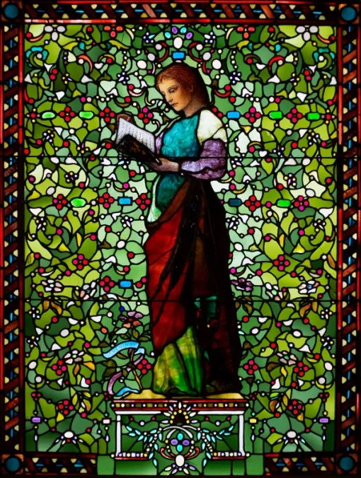Vibrant stained glass window with a woman shown standing at center and looking downwards and to the left at an open book that she holds in her hands. She is surrounded by abstract forms that evoke leaves and flowers and a decorative frame.