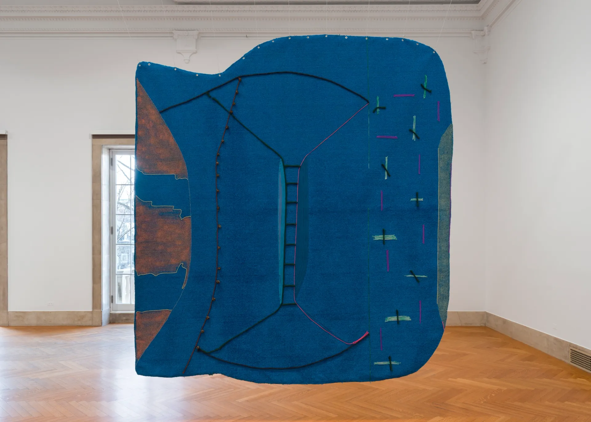 Large suspended painting in a gallery space; blue astroturf delineated with colorful yarn and paint; seed pods stitched to surface