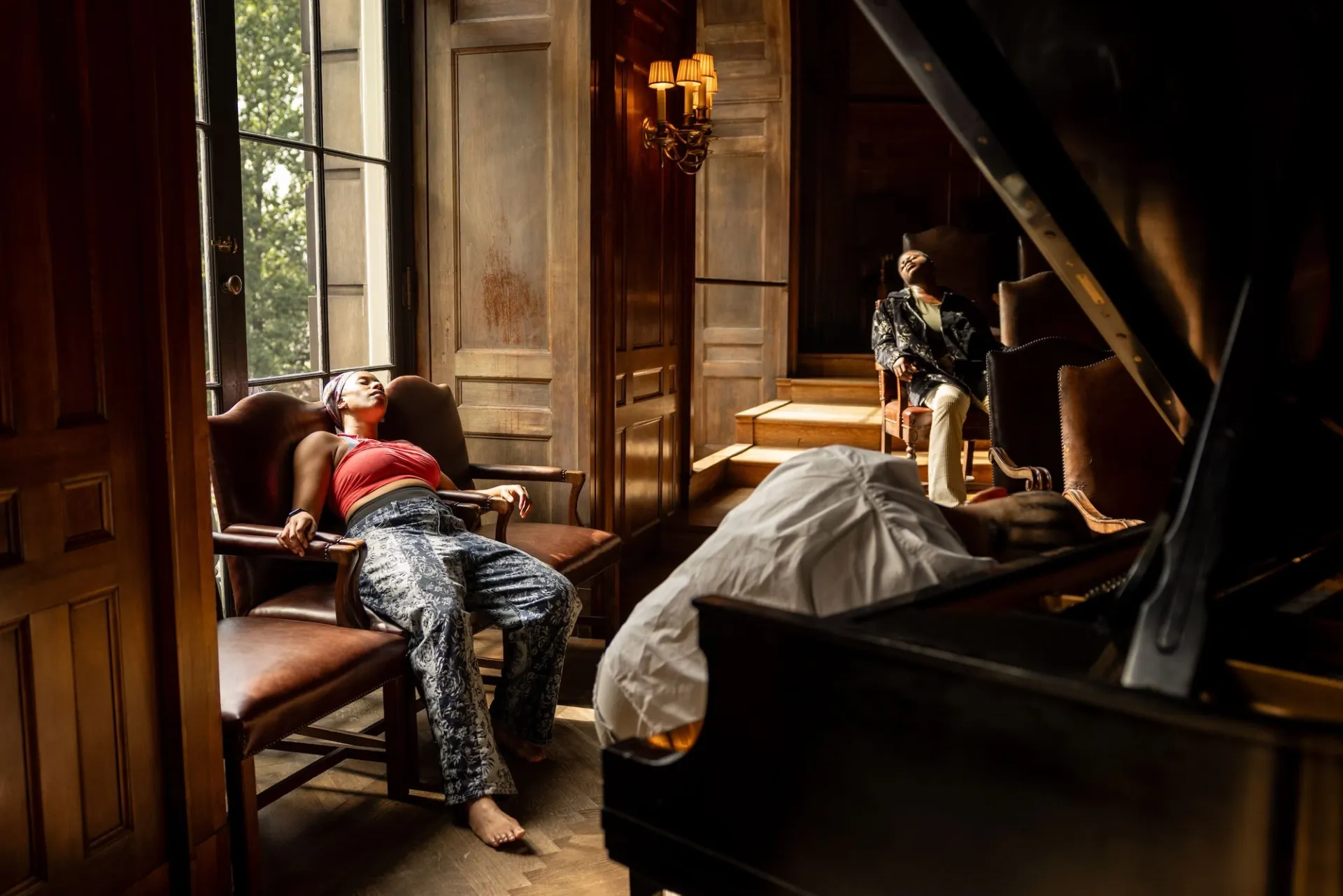 Three performers recline in a wood-paneled space with leather chairs and large windows; one leans onto the keyboard of a grand piano