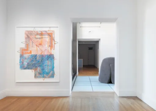 On a wall to the left of an entryway hangs a framed drawing with blue, orange, and peach geometric forms. Beyond the entryway, a soft, gray sculpture rests on the floor in the adjoining room.