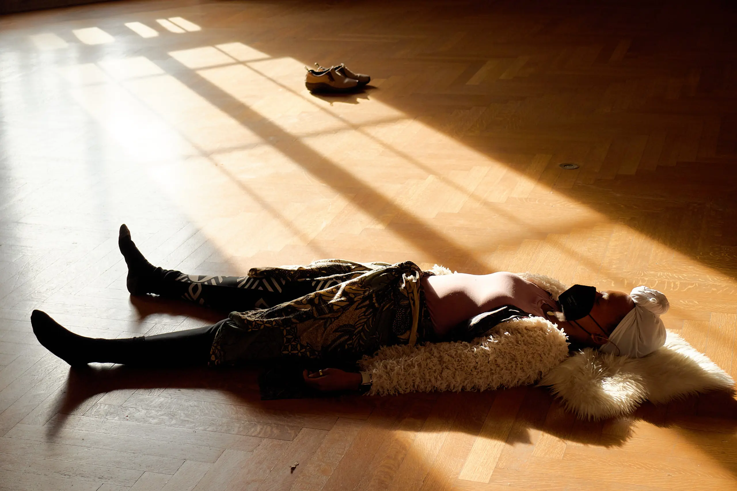 A person with eyes closed lying on her back on a wooden floor; sunlight streams into the room from nearby window