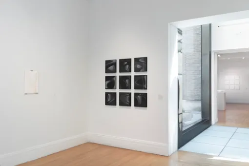 At an angled view, a single art piece hangs on the wall to the left. Adjacent to it lies a series of squares composing a single artwork. To the right, an entryway with a a large window leads to another gallery with artworks visible inside.