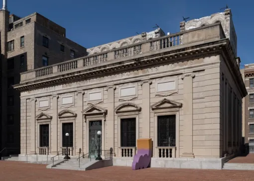 An exterior view American Academy of Arts and Letters shows the building’s neoclassical architecture. To the right of the entrance, a large two-toned installation piece leans against the facade, nestled between two windows.