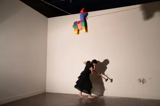 A barefoot woman in a dress takes a sledgehammer to a wall, her body casting a large shadow, as a colorful piñata sits suspended by string in the air above her