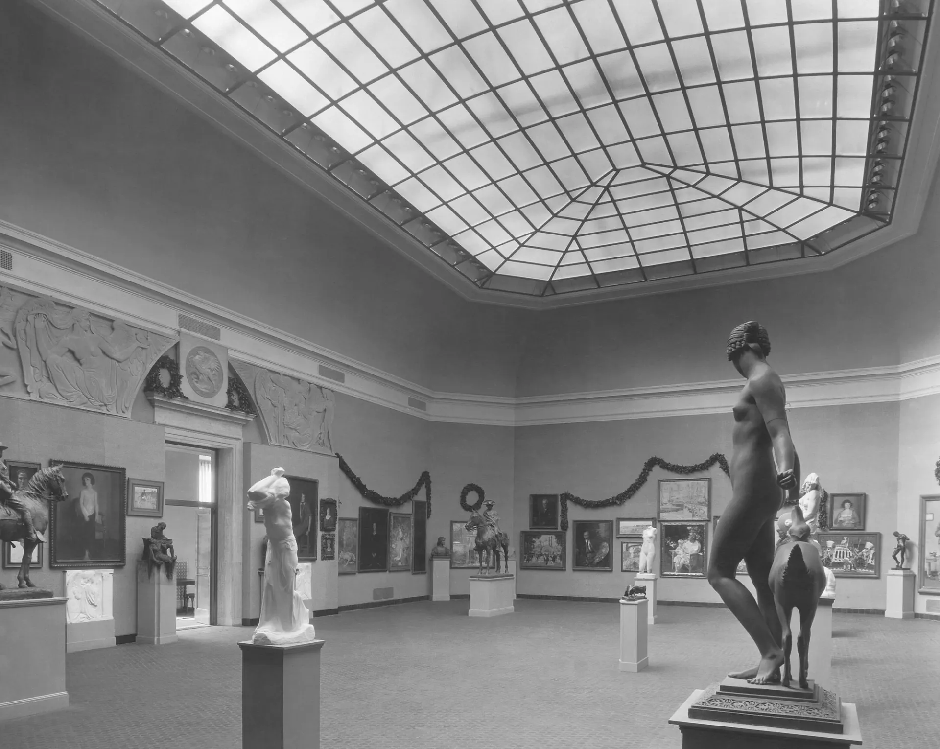 Sculptures and paintings installed in a large gallery whose ceiling is a skylight. Garlands of greenery also adorn the walls.