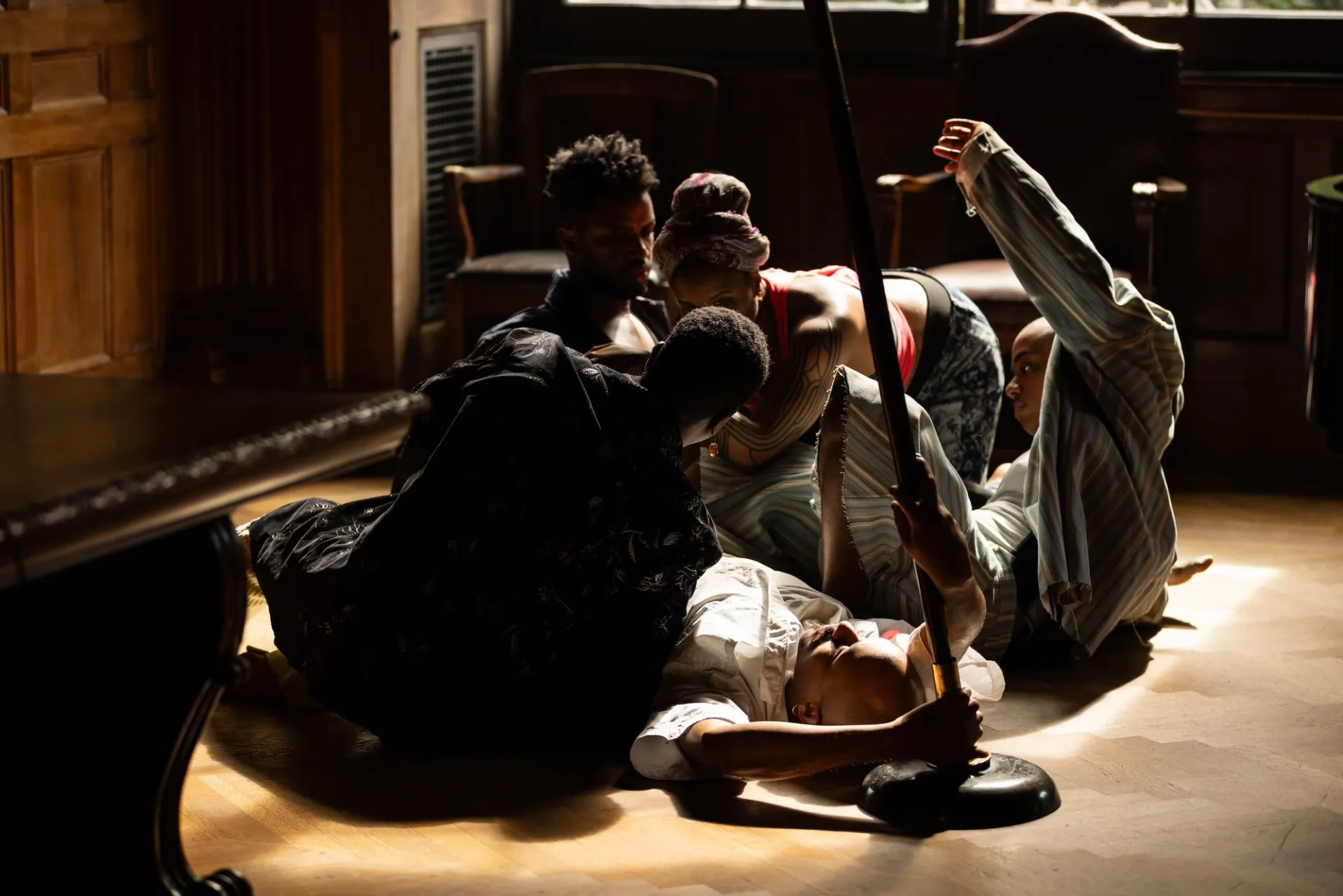 A group of performers cluster together in a sunny spot on a wooden floor