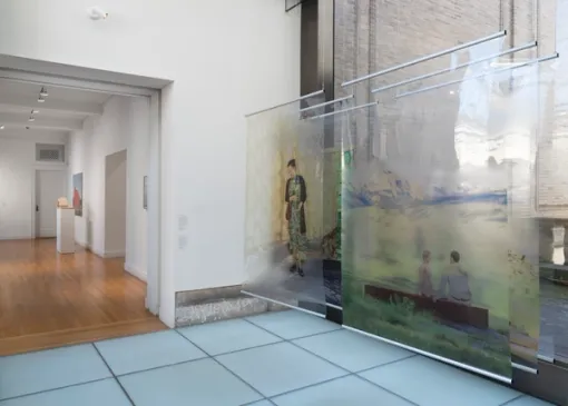 A series of large floor-to-ceiling artworks on transparent scrolls hang suspended to the right. To the left lies another gallery with artworks on the wall and an object on a pedestal. 