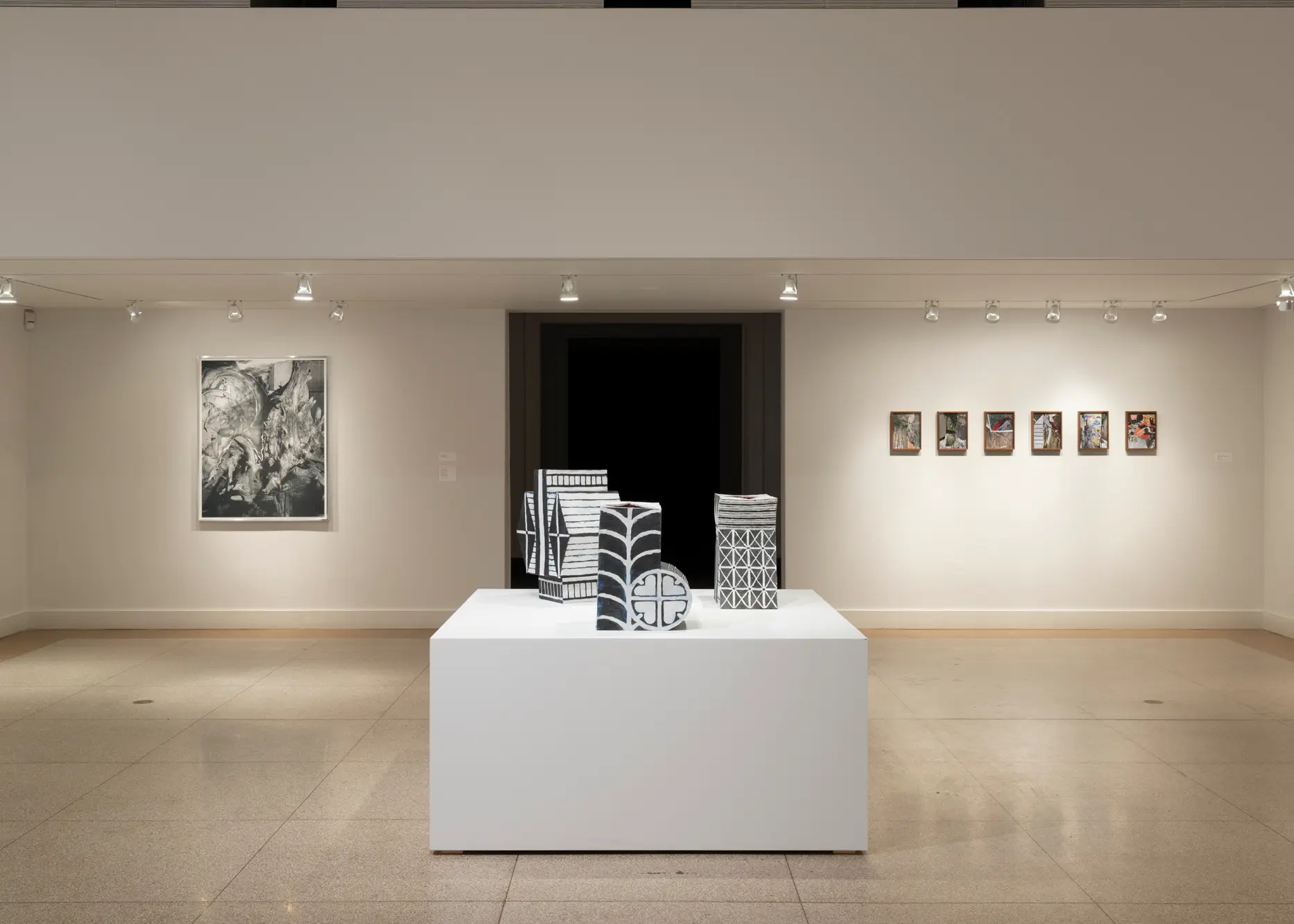 Three black and white sculptures sit on a large white pedestal at the center of a gallery room. On the wall behind the sculptures, a large abstract work on paper is hung to the left of a series of six small, framed works.