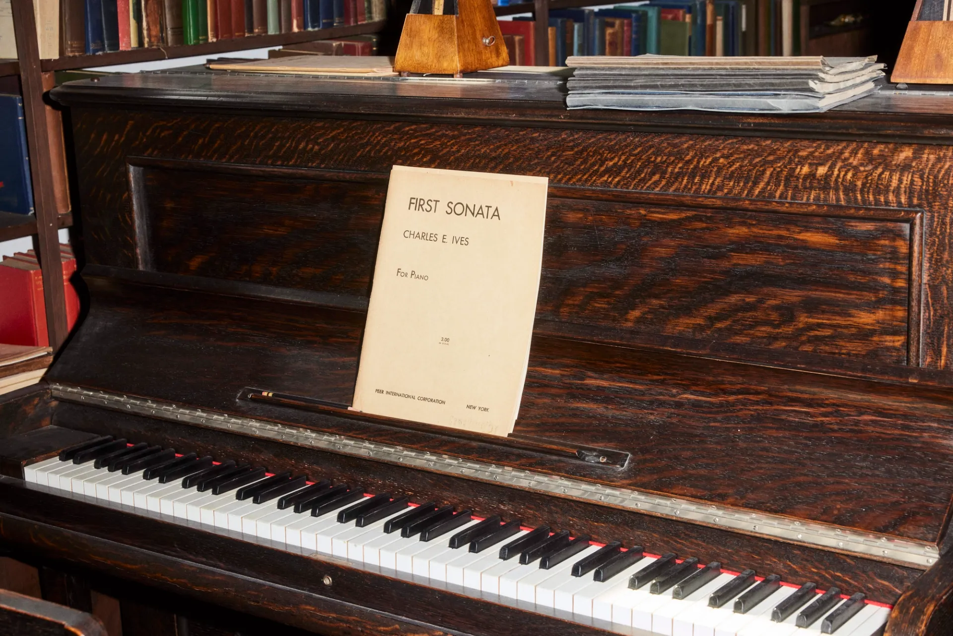 A dark wooden upright piano with a piece of sheet music on it attributed to Charles Ives.