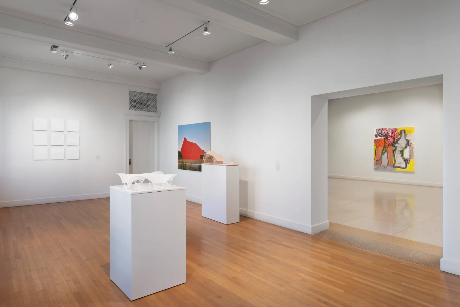Architectural models sit on plinths in the center of a room and against a wall. To the left, art in a series of colorless squares comprise a single artwork. Against the adjacent wall lies a colorful artwork. To the right, and through an entryway, a single painting adorns a wall