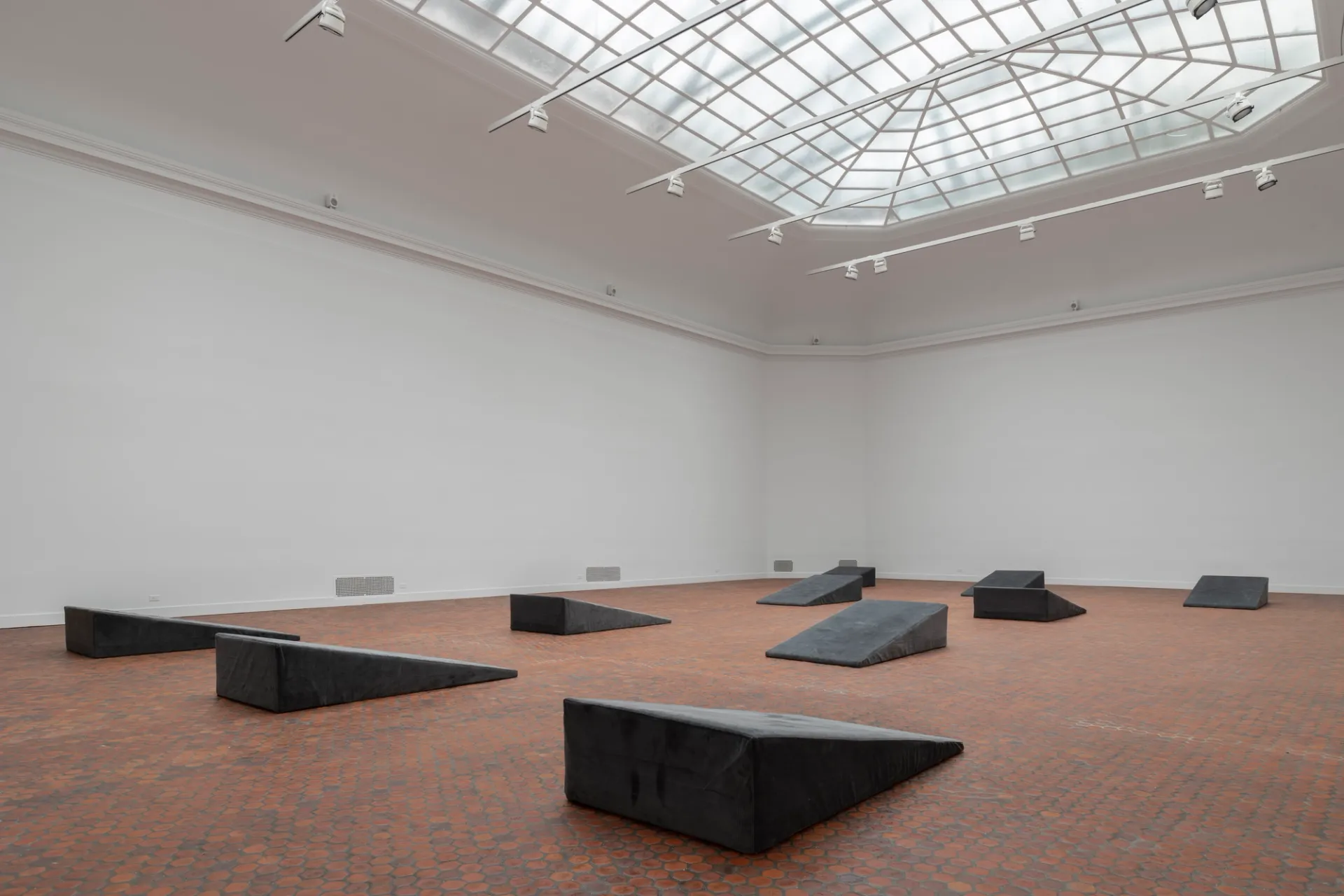 A skylit gallery with ten wedge shaped cushions on the floor.
