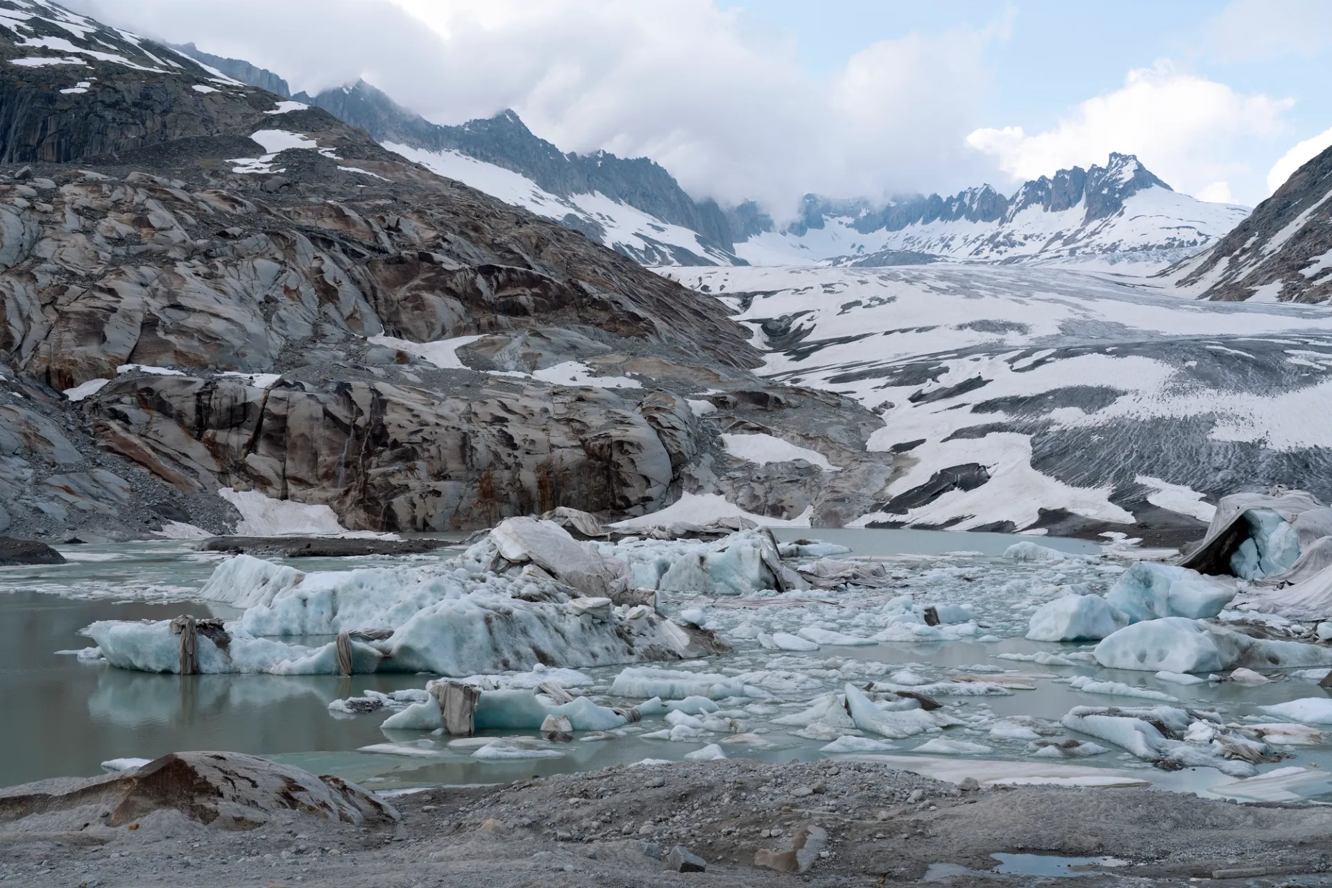 Photograph of a glacier, partially covered with blankets, with water in the foreground. Snow-capped mountains recede into clouds in the background.