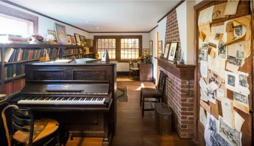 A long room contains a wall-to-wall bookshelf filled with scores, a piano, chairs, photo frames, and clippings pinned to a wooden door.