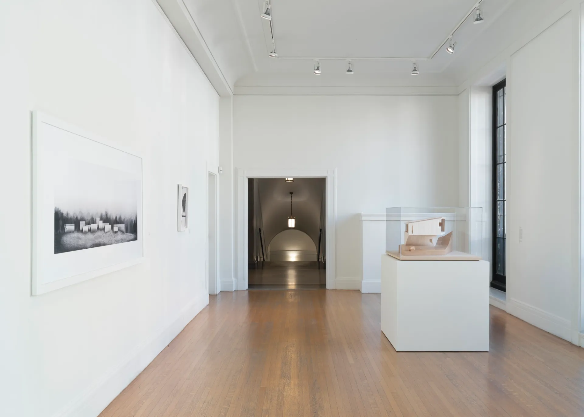 To the left, framed artwork hangs on the wall. Directly ahead, an entryway opens onto a staircase. To the right, an architectural model encased in plexi sits on a pedestal. 