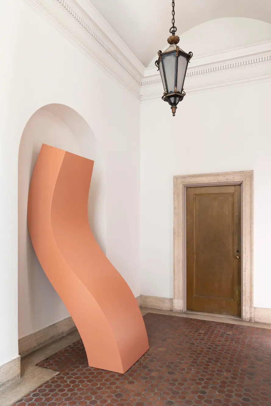 In a gallery space with spanish tile floor, a salmon colored rectangular sculpture with a tall, undulating form leans against the wall.