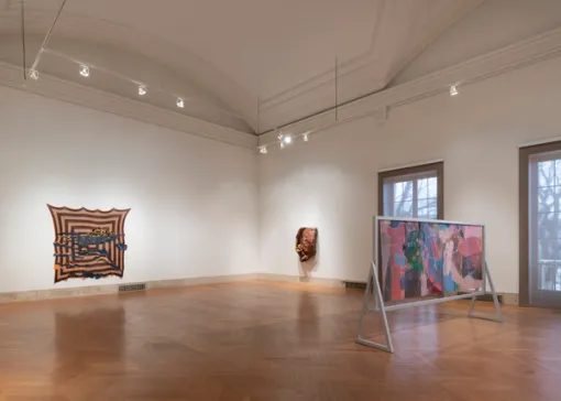 A large colorful painting mounted in a wooden armature stands in the gallery before a geometric, fabric work and abstract sculpture hanging on the adjacent walls.