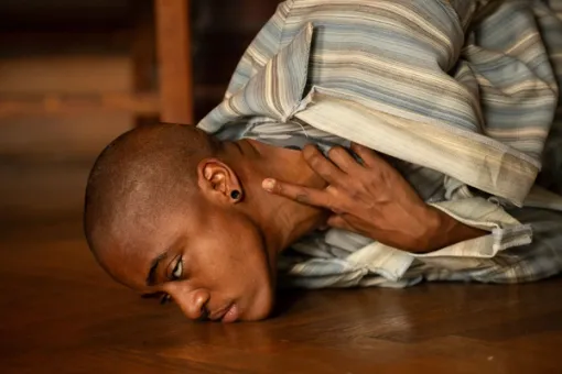 Close-up of a performer seen in profile; their lips and chin rests on a wooden floor and their hand holds their throat