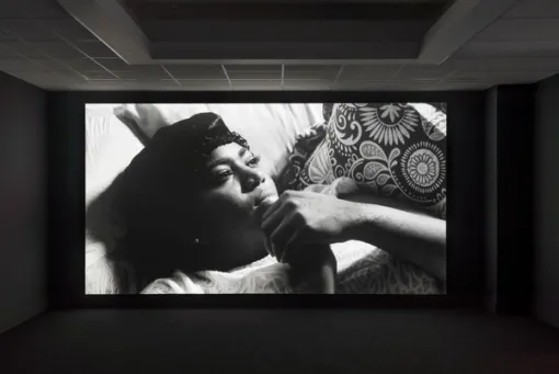 An image of a darkened theater with a black and while film still of a woman on her back looking up and clasping her hands together. 