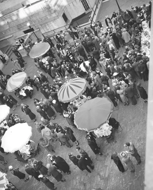 A black and white photograph shows a celebration on the Audubon Terrace from above. Within the skewed image, groups stand amidst round tables with decorative umbrellas.